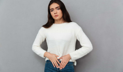 Close-up photo of depressed young woman with abdominal pain, looking at camera, isolated over gray background